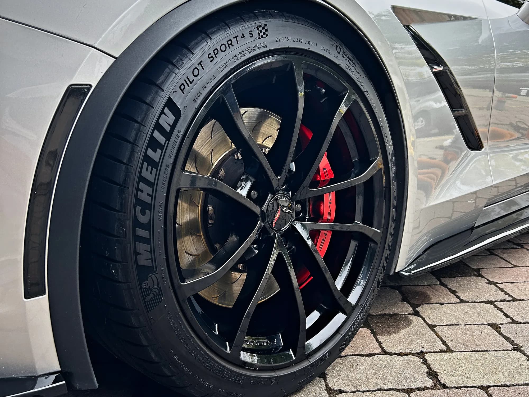 A Corvette Grand Sport wheel after detailing: gloss black spokes in front of a red caliper, the tire dressed black, silver paint reflecting the sky