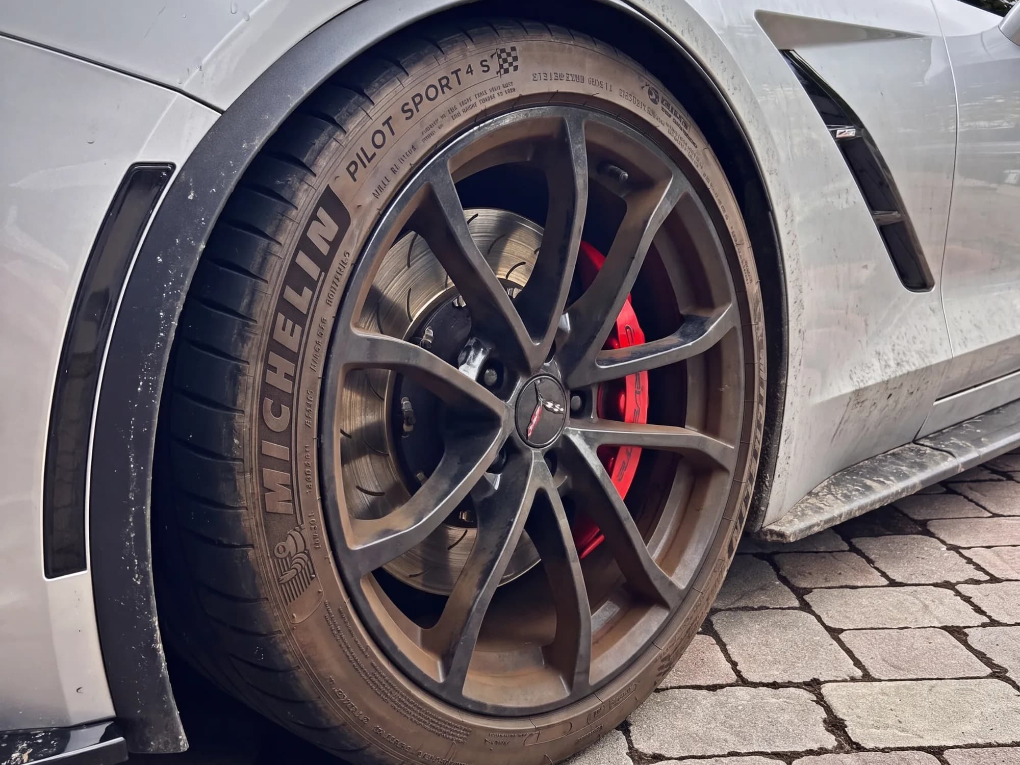 The same Corvette Grand Sport wheel, recreated from the after photo to show it before cleaning: a dust film over the silver paint, brown brake dust on the black spokes, and a faded tire sidewall